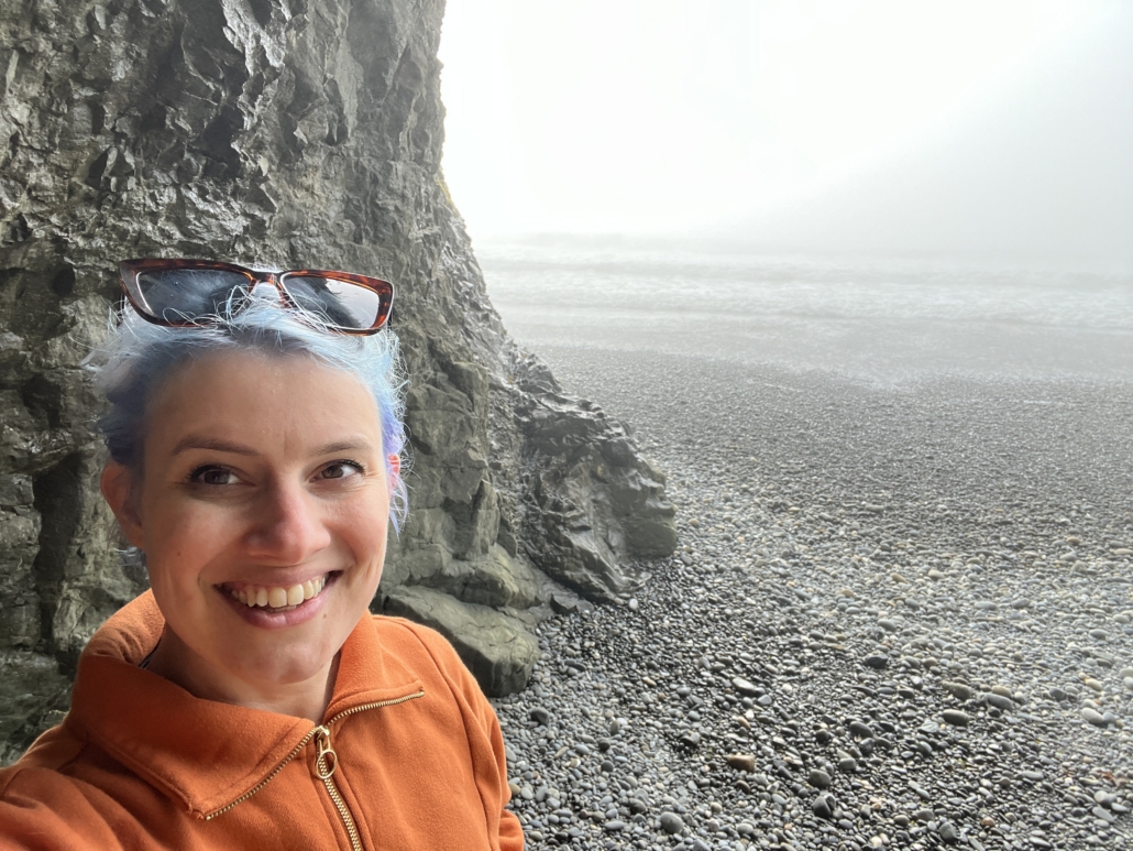A white woman stands next to a rock wall on a foggy day at a beach on the Pacific Ocean.